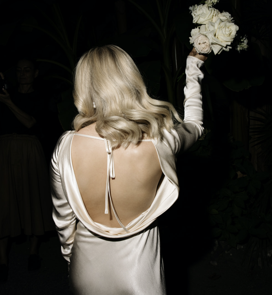 Nærbilde av ryggen til en brud i en hvit kjole som holder en bukett under et overraskelsesutdrikningslag.